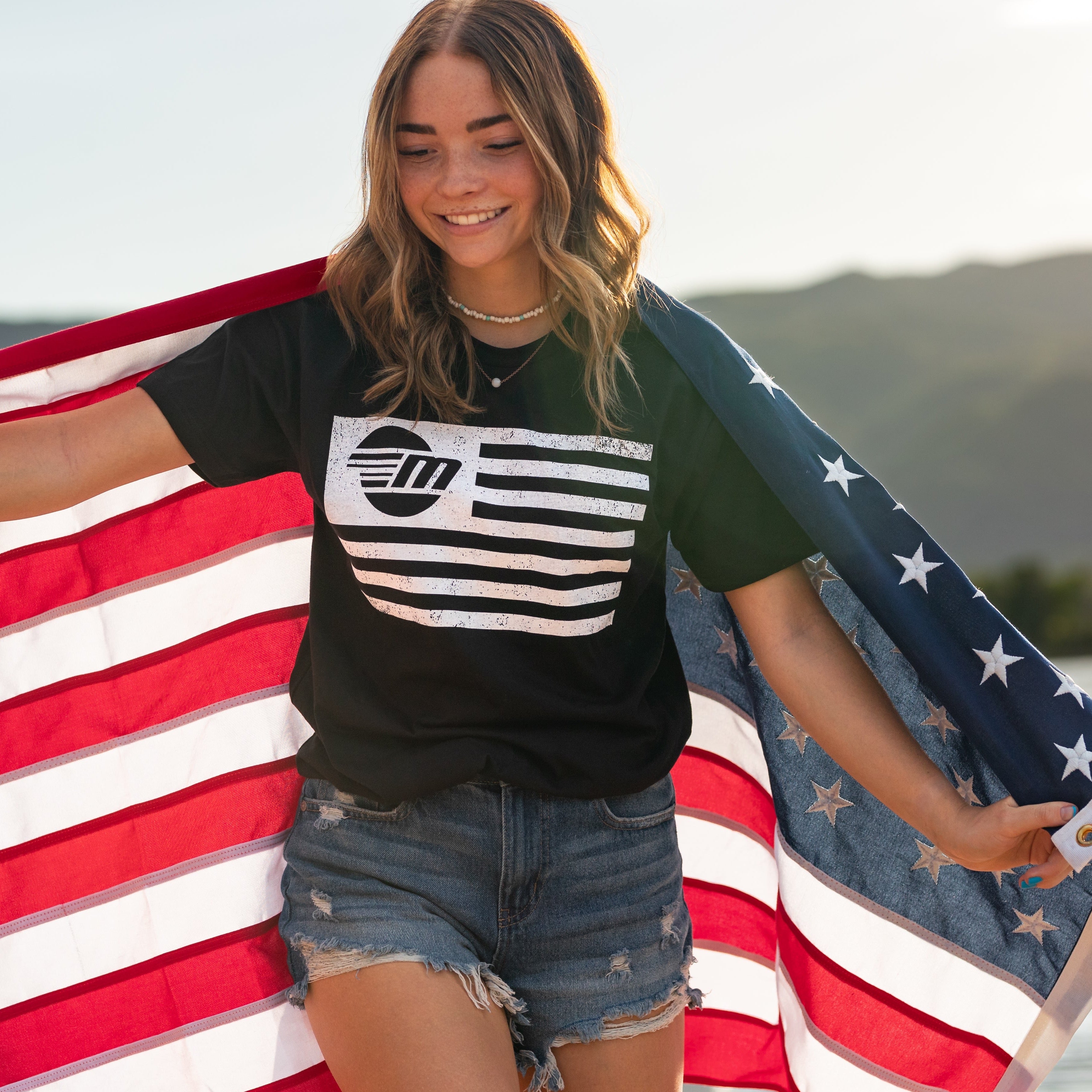Young woman smiling outdoors, holding an American flag, wearing a black Malibu Freedom Tee with patriotic design.