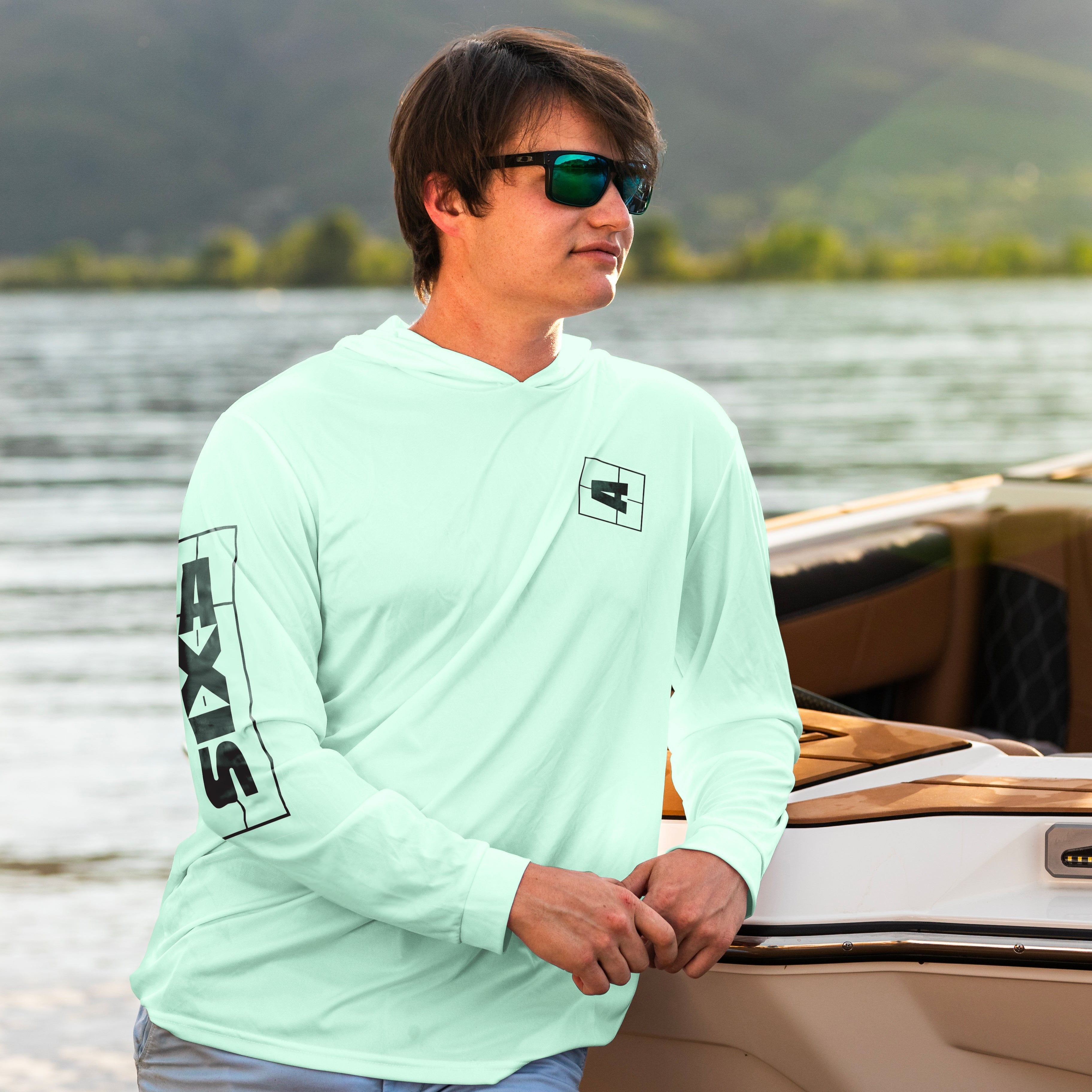 Man wearing a light green shaded shredder sun hoodie with AXIS logo, standing by a boat on a lake with mountains in the background.