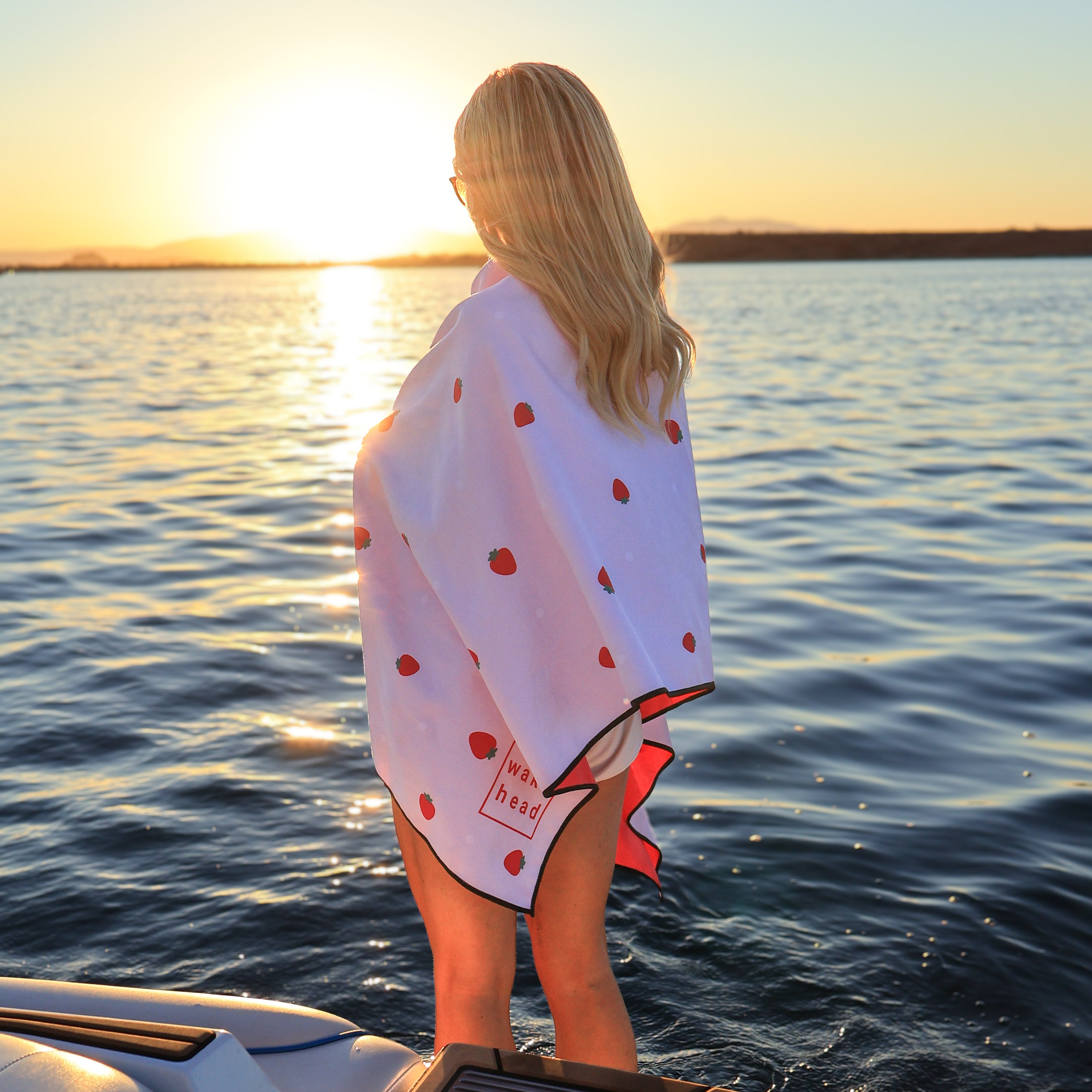 Woman with blonde hair wrapped in a white berry-themed lake towel, standing on a boat at sunset over a calm lake.
