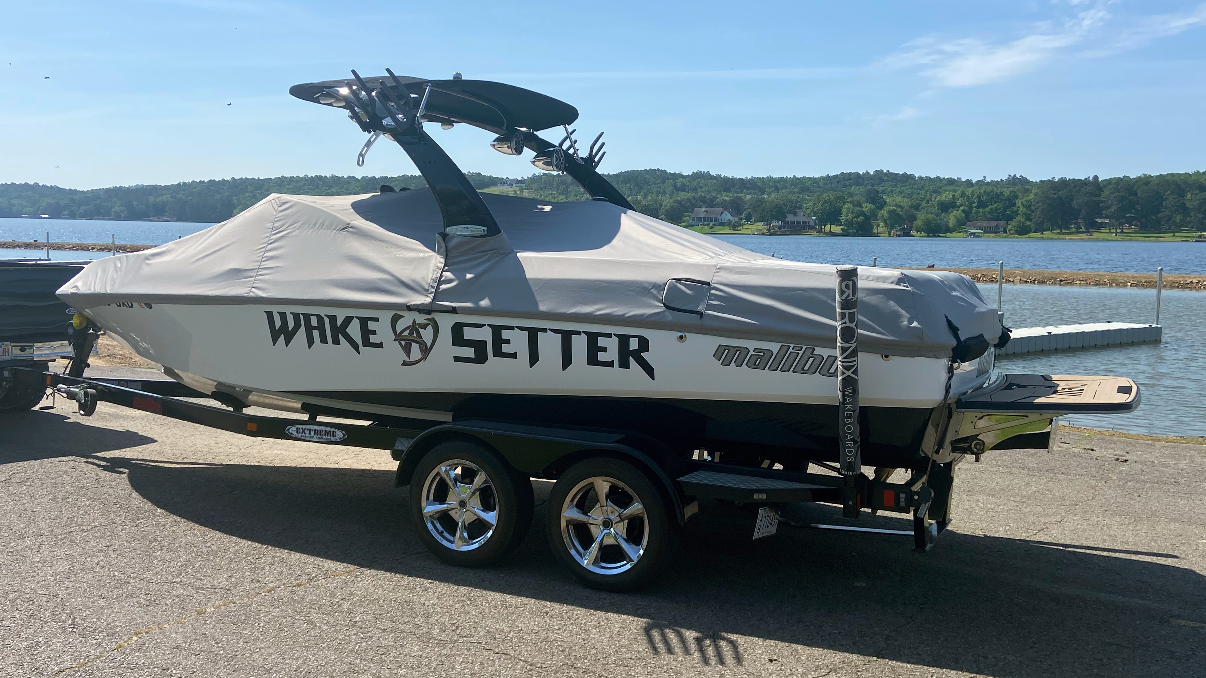 Boat covered with a Sunscape/Wakesetter 247 cover, mounted on a trailer with Illusion X/XS tower, parked near a lake.