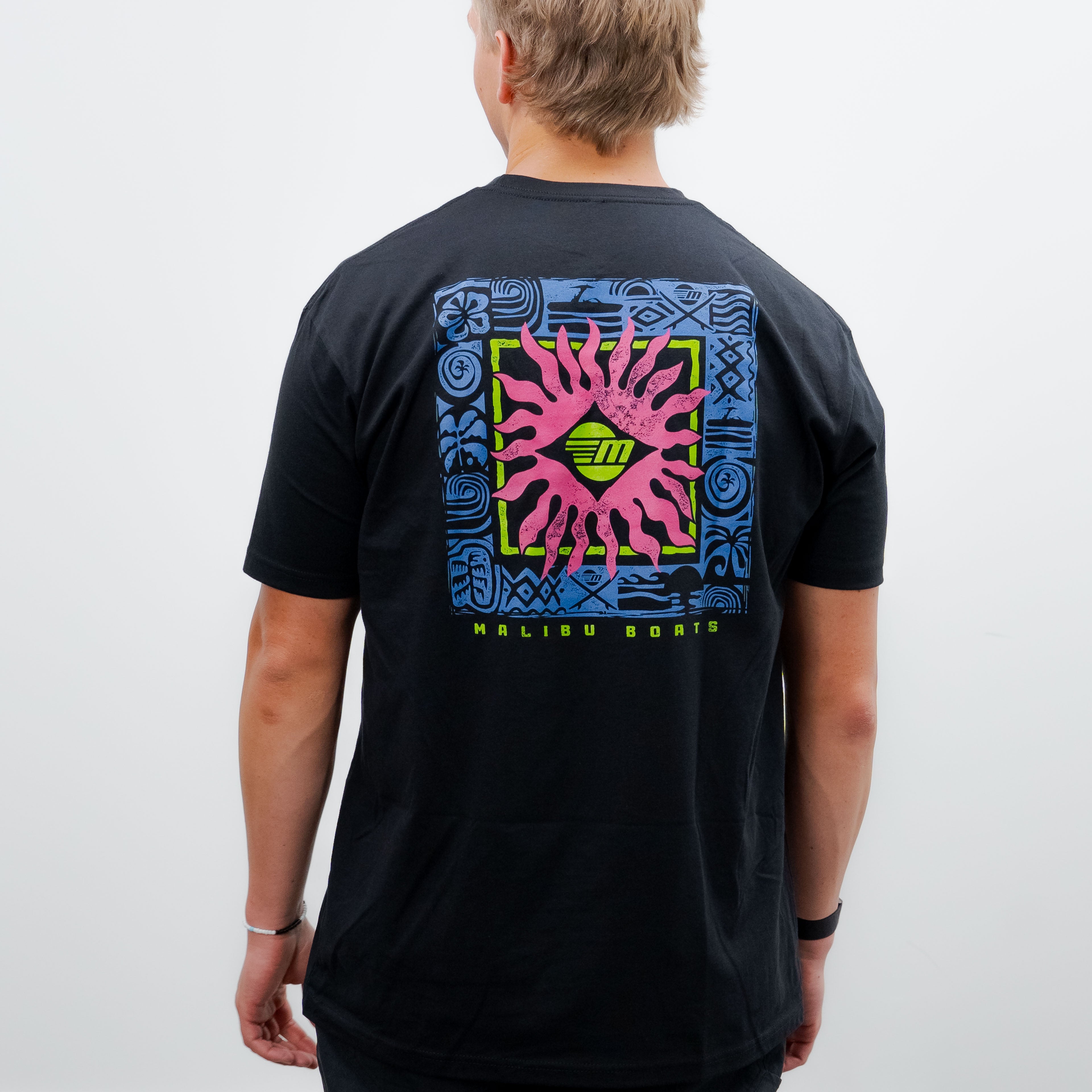Back view of a person wearing a black Malibu Tribal Tee with colorful tribal-inspired graphic design and "Malibu Boats" text.