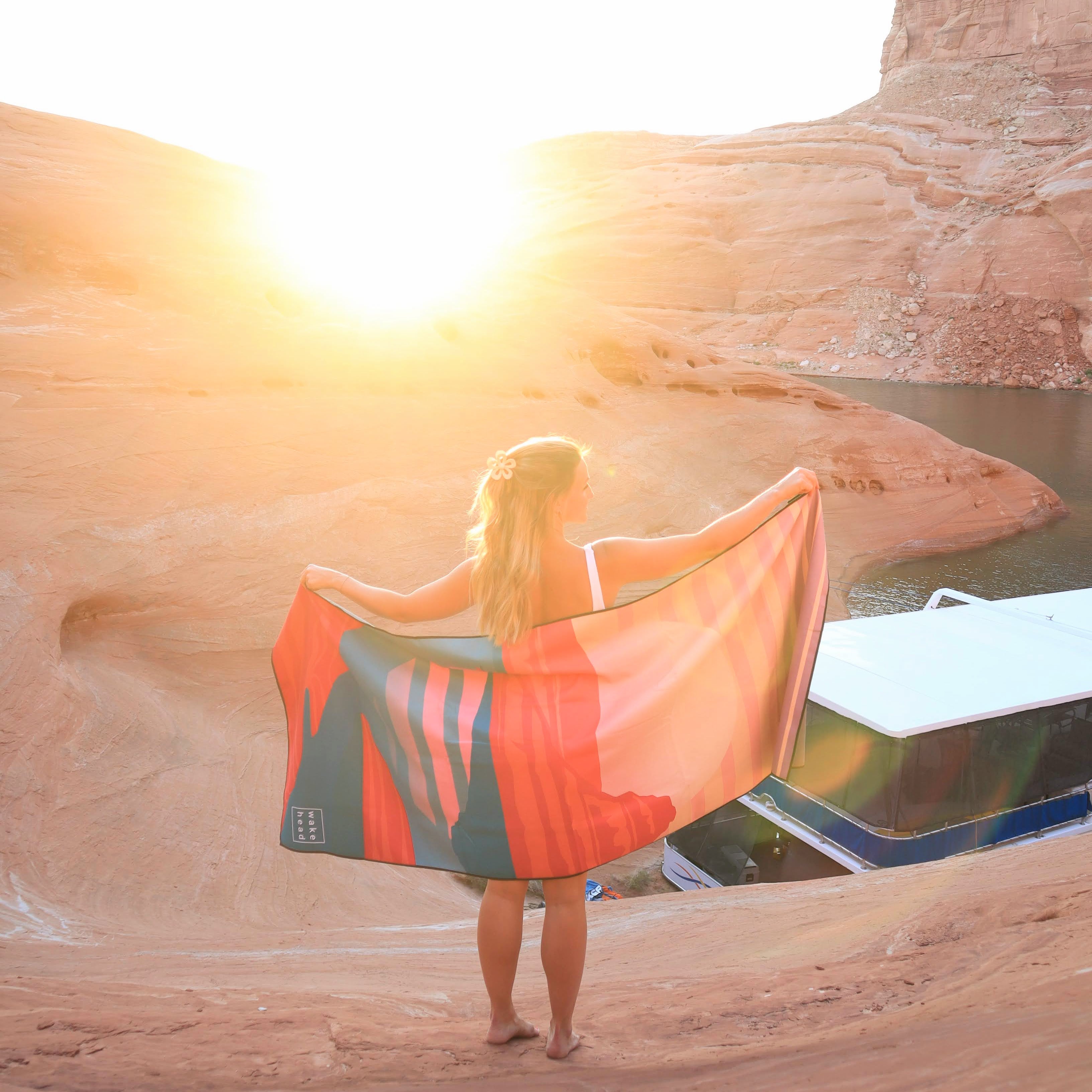 Woman holding a Powell Towel at sunset near a desert canyon with water, showcasing the product's vibrant design and outdoor use.
