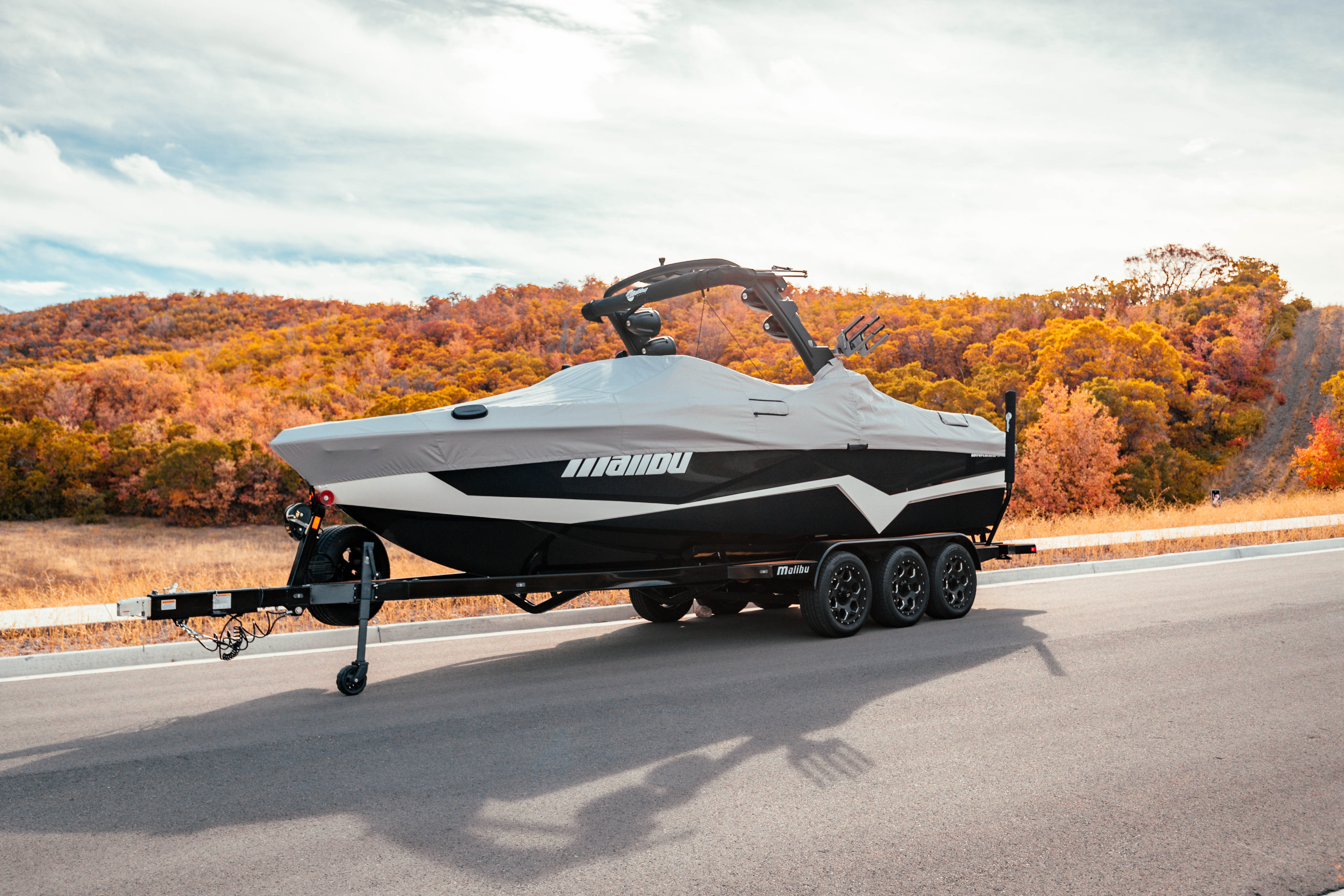 A Malibu boat on a trailer with a 23 LSV cover, bow cleats, and G3 tower, parked on a road with autumn foliage in the background.